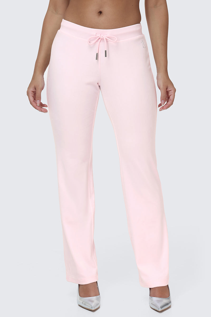 A person wearing the OG Big Bling Velour Track Pants in light pink, featuring a subtle Juicy embellishment, paired with silver pointed-toe heels and standing against a plain light gray background.