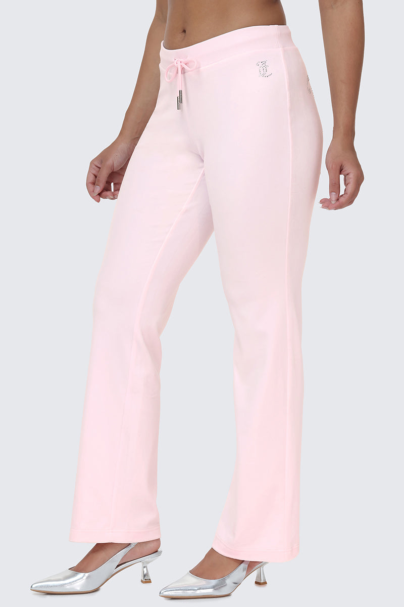 A person wearing light pink OG Big Bling Velour Track Pants with a flared silhouette and drawstring waist, paired with silver high-heeled shoes, stands against a plain gray background. The subtle Juicy embellishment on the pants adds a touch of playful Y2K nostalgia to the look.