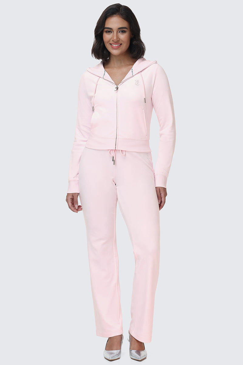 A woman stands and smiles on a plain light background, wearing the OG Big Bling Velour Track Pants with matching light pink velour zip-up hoodie featuring Juicy embellishment, paired with silver pointed-toe shoes.