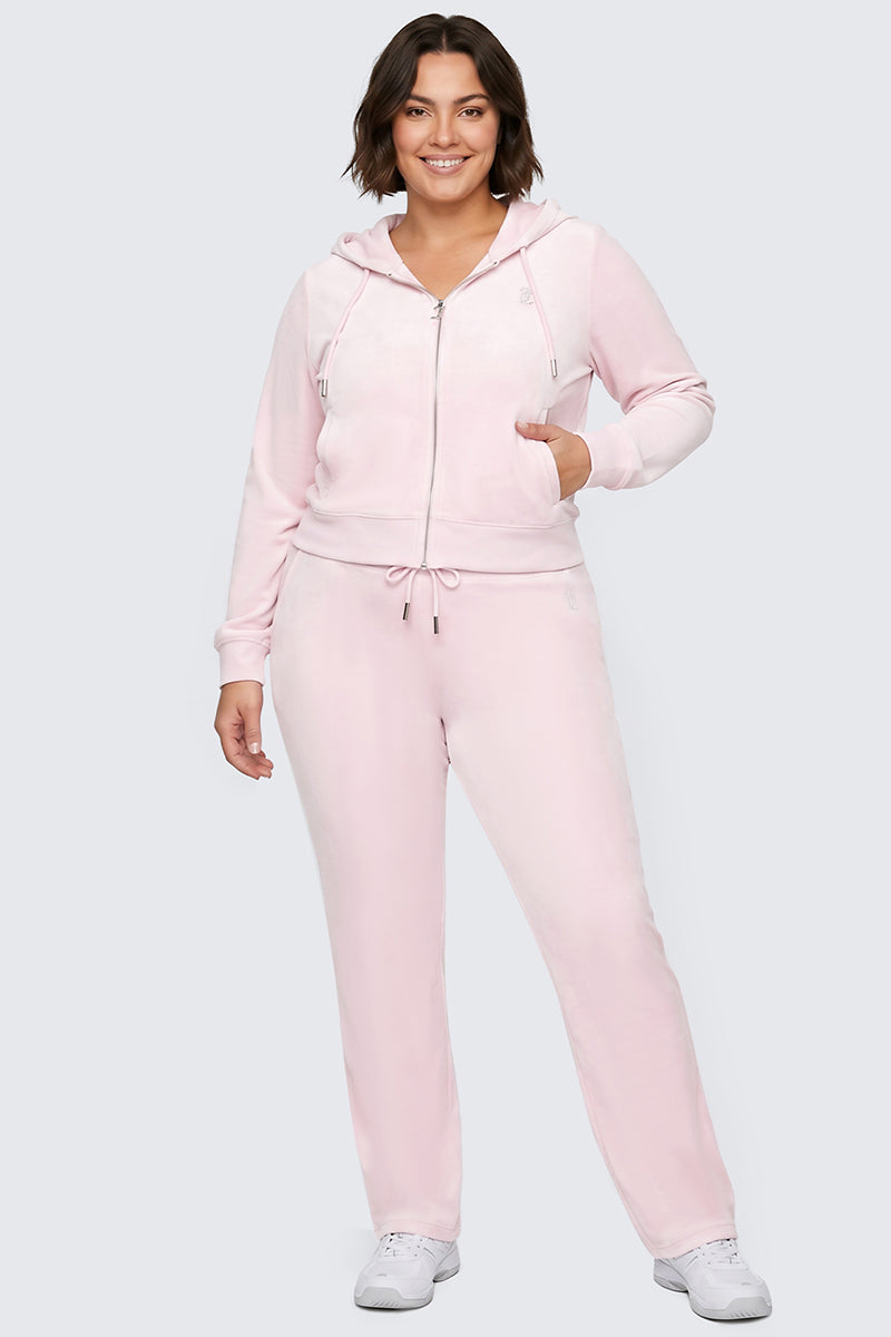 A woman with short brown hair wears a light pink zip-up hoodie and the Plus-Size OG Big Bling Velour Track Pants, paired with white sneakers, standing and smiling against a plain light gray background.