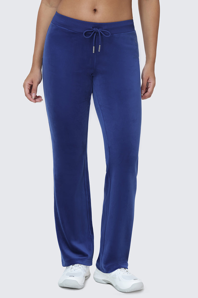 A person wearing OG Big Bling Velour Track Pants in blue, featuring subtle Juicy embellishment and a drawstring waist, paired with white sneakers. Only the lower half of the body is visible against a plain light background.