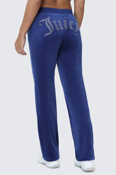 A person is wearing the OG Big Bling Velour Track Pants, featuring Juicy embellishment in rhinestones on the back, standing and facing away from the camera. White sneakers complete this iconic Y2K look.