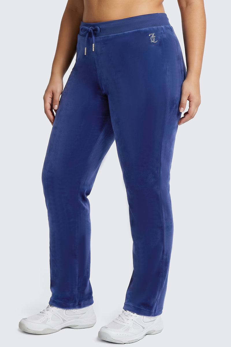 A person is shown from the waist down, wearing Plus-Size OG Big Bling Velour Track Pants in blue with a subtle Y2K flair and white sneakers, standing against a plain light gray background.