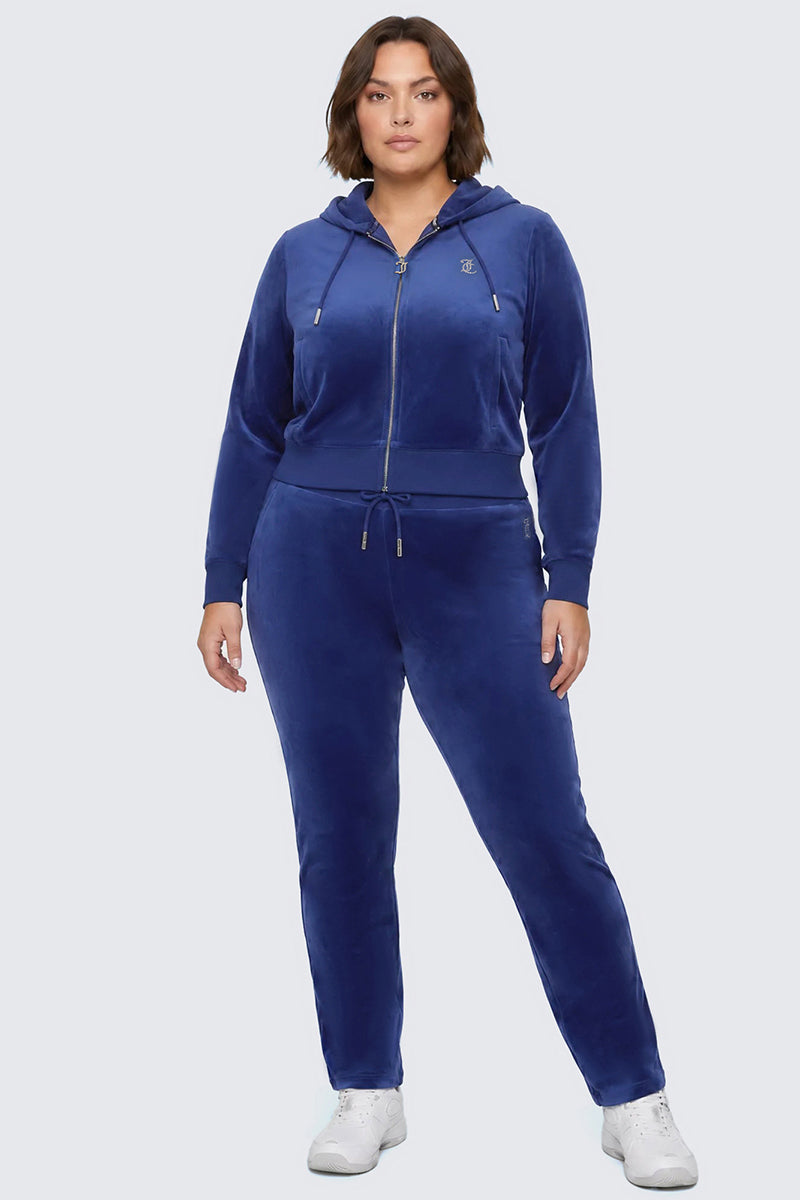 A woman stands facing forward against a light gray background, wearing the Plus-Size OG Big Bling Velour Track Pants in royal blue with classic Y2K vibes—paired with a matching zip-up hoodie and white sneakers.