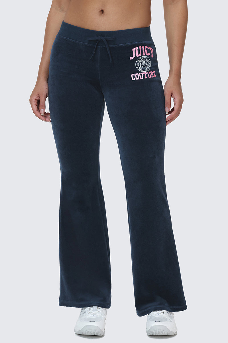 A person wearing Scottie Cotton Velour Track Pants in navy blue, featuring "Juicy Couture" and a logo in pink and white on the left hip, channels iconic Y2K style paired with white sneakers; the snap back pocket pants stand out against a light gray background.