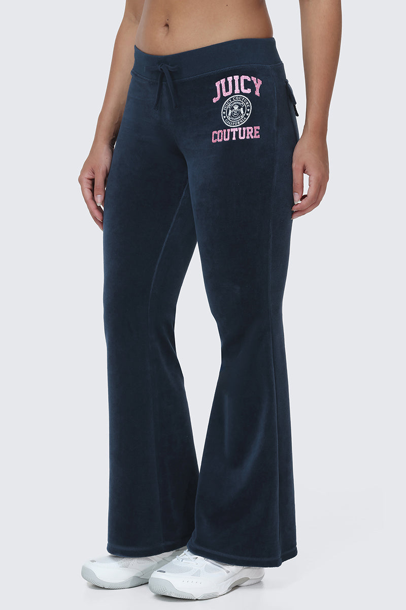 A person wearing Scottie Cotton Velour Track Pants in dark blue, featuring pink and white Juicy Couture crest logos on the left hip for a classic Y2K vibe, paired with white sneakers.