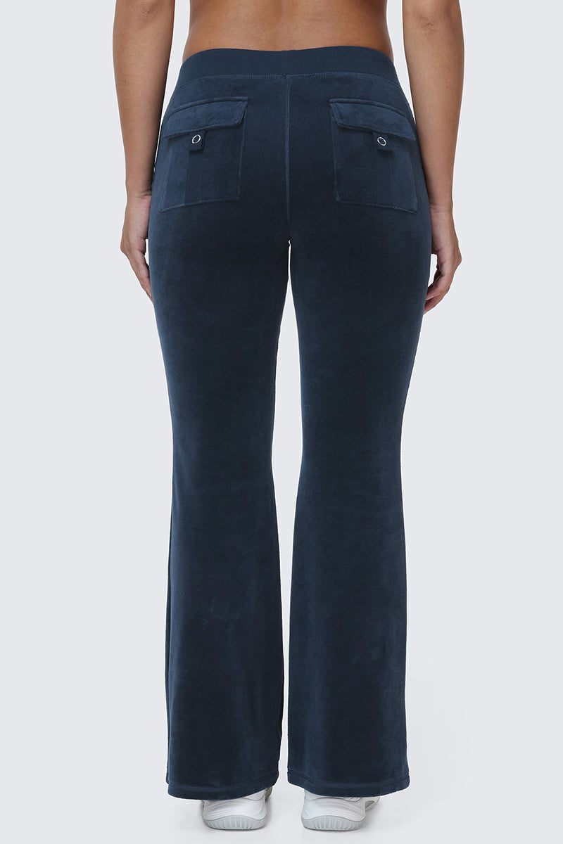 A person is seen from behind wearing the Scottie Cotton Velour Track Pants in dark blue, which feature snap back pockets and a slight flare at the bottom, channeling Y2K style. The look is completed with white shoes.