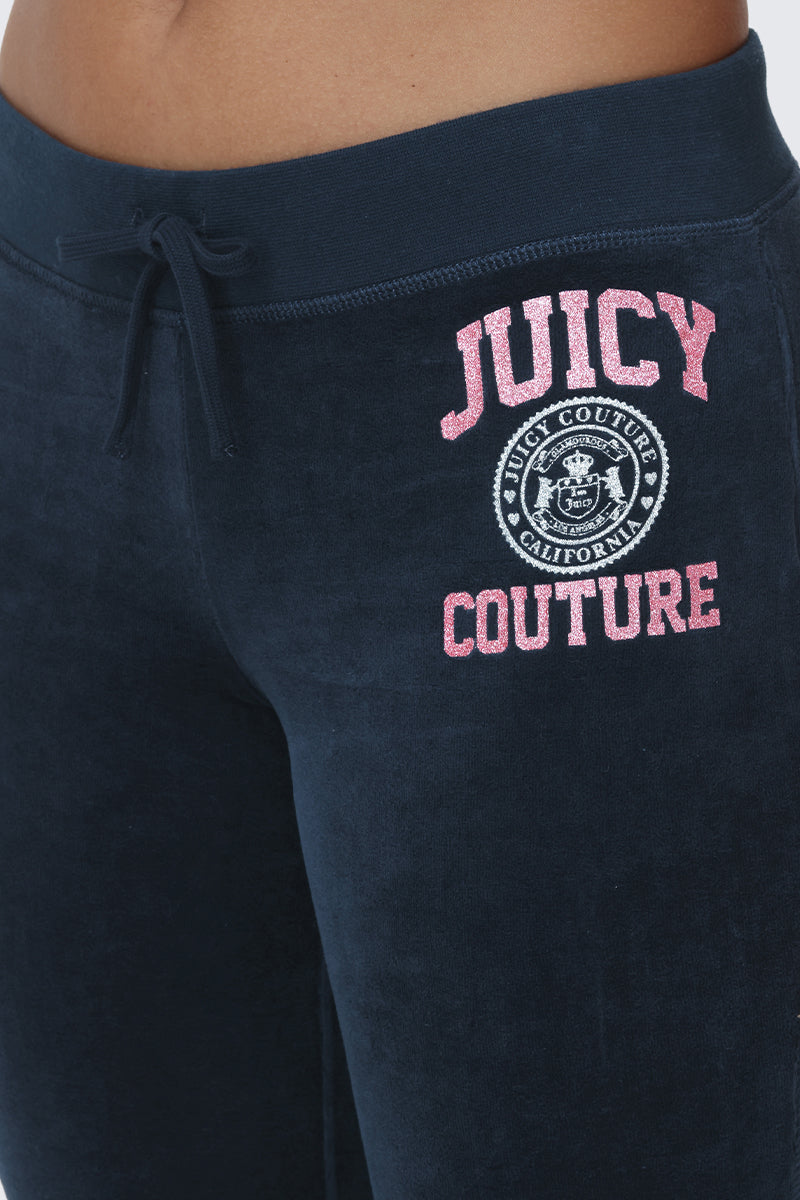 Close-up of a person wearing navy blue Scottie Cotton Velour Track Pants by Juicy Couture, featuring pink JUICY COUTURE text and a white circular logo on the left hip, channeling true Y2K style.