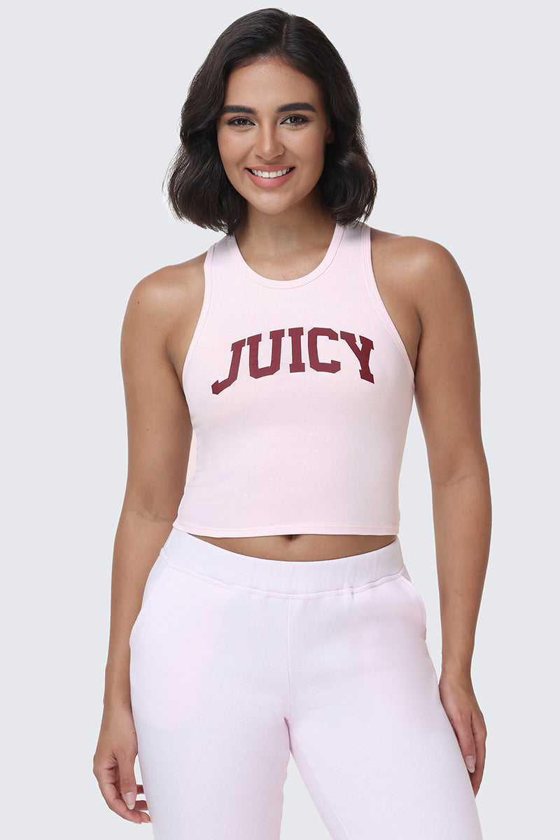 A woman with shoulder-length dark hair wears the Collegiate Juicy High Neck Tank Top in light pink, featuring a bold red JUICY logo in a collegiate style. She pairs it with matching light pink pants, standing against a plain light gray background and smiling.