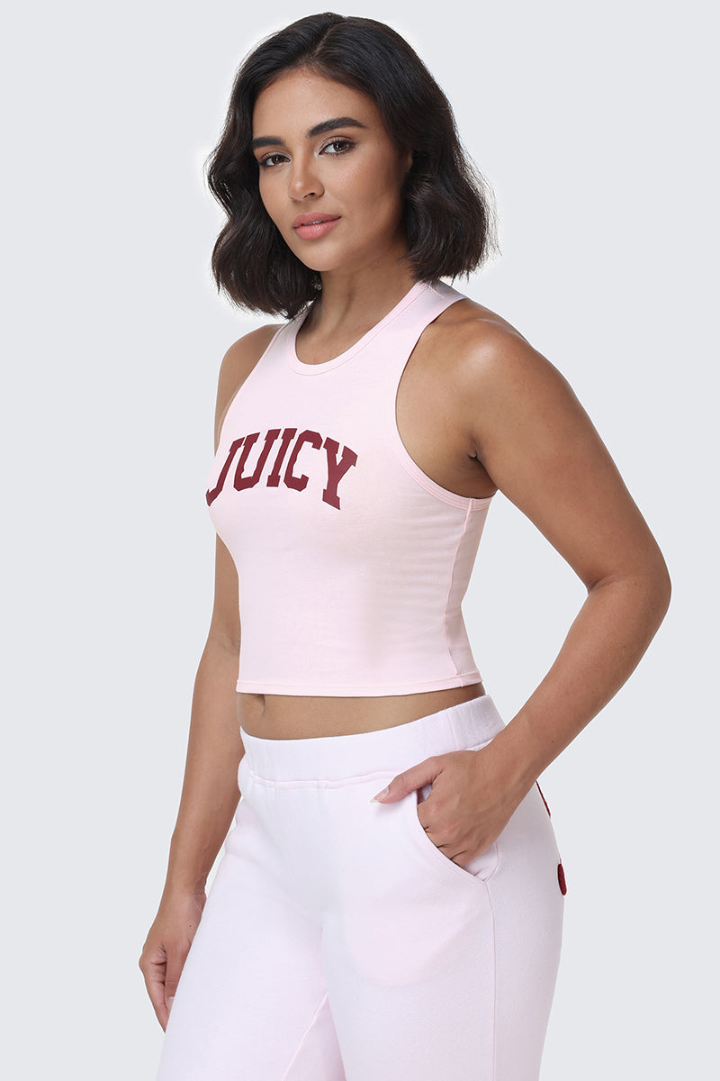 A woman with short dark hair is wearing the Collegiate Juicy High Neck Tank Top in light pink, featuring a bold red Juicy logo in collegiate style, paired with matching light pink pants. She poses against a plain background with one hand in her pocket.