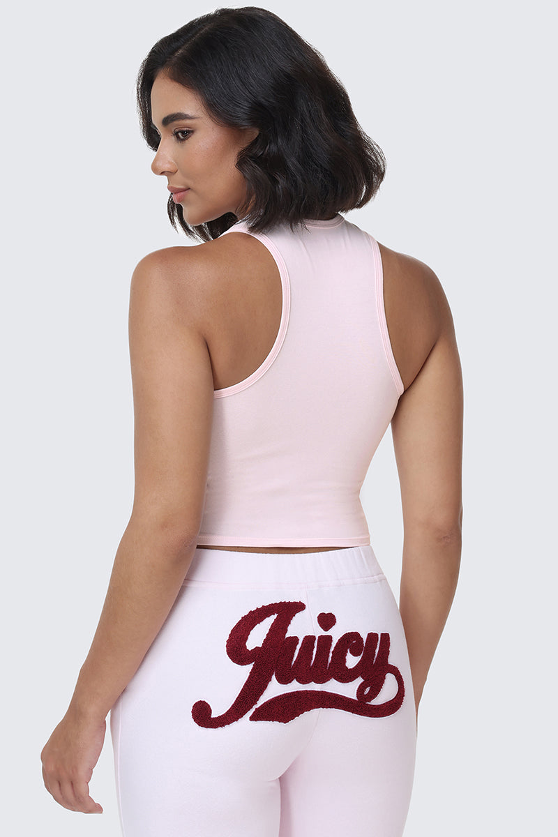 A woman with wavy dark hair wears the Collegiate Juicy High Neck Tank Top in light pink and matching pants featuring the Juicy logo in red script on the back, standing against a plain gray background.