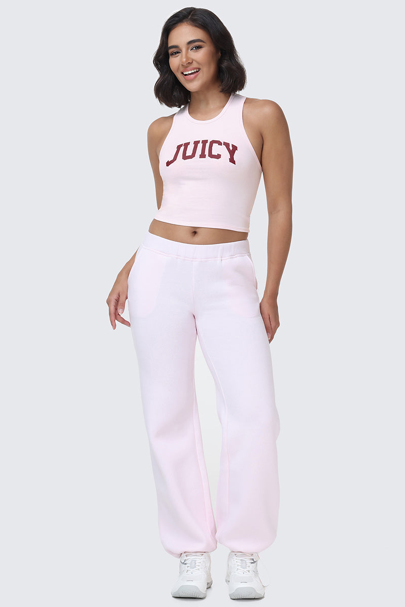 A woman is standing and smiling, wearing the Collegiate Juicy High Neck Tank Top in light pink with a bold red Juicy logo, paired with matching light pink sweatpants and white sneakers.