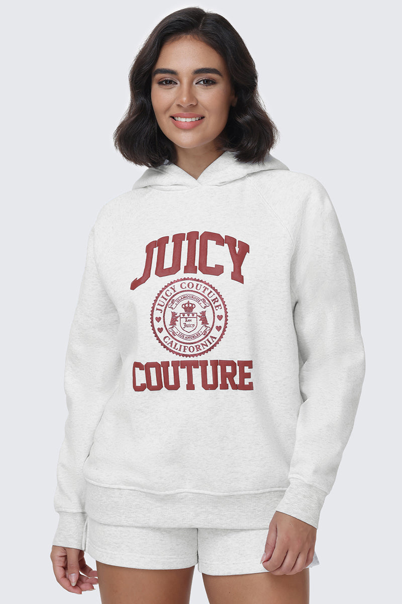 A woman stands smiling against a plain light background, wearing the Scottie Fleece Oversized Hoodie in white and matching shorts, featuring "Juicy Couture California" in red text and a bold Juicy crest.