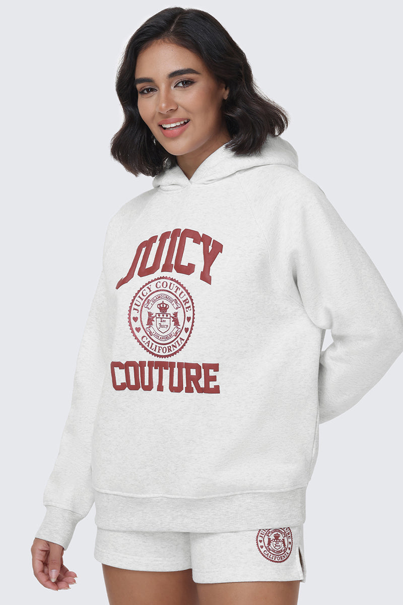 A woman smiles while wearing the Scottie Fleece Oversized Hoodie and matching shorts set in white, featuring bold red "Juicy Couture California" lettering and the iconic Juicy crest on both the hoodie and shorts, as she stands against a plain light gray background.