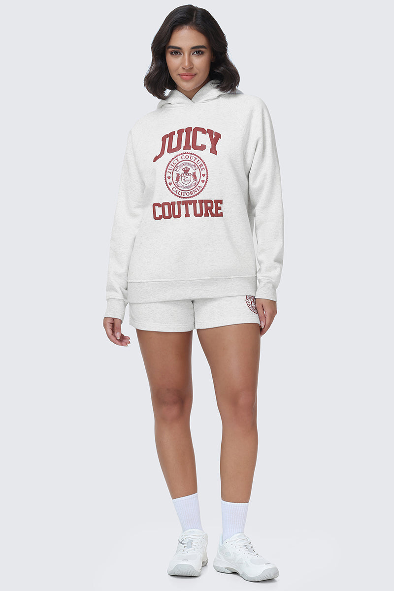 A woman stands and smiles, wearing the light gray Scottie Fleece Oversized Hoodie by Juicy Couture with matching shorts featuring red lettering, paired with white socks and white sneakers against a plain light background.