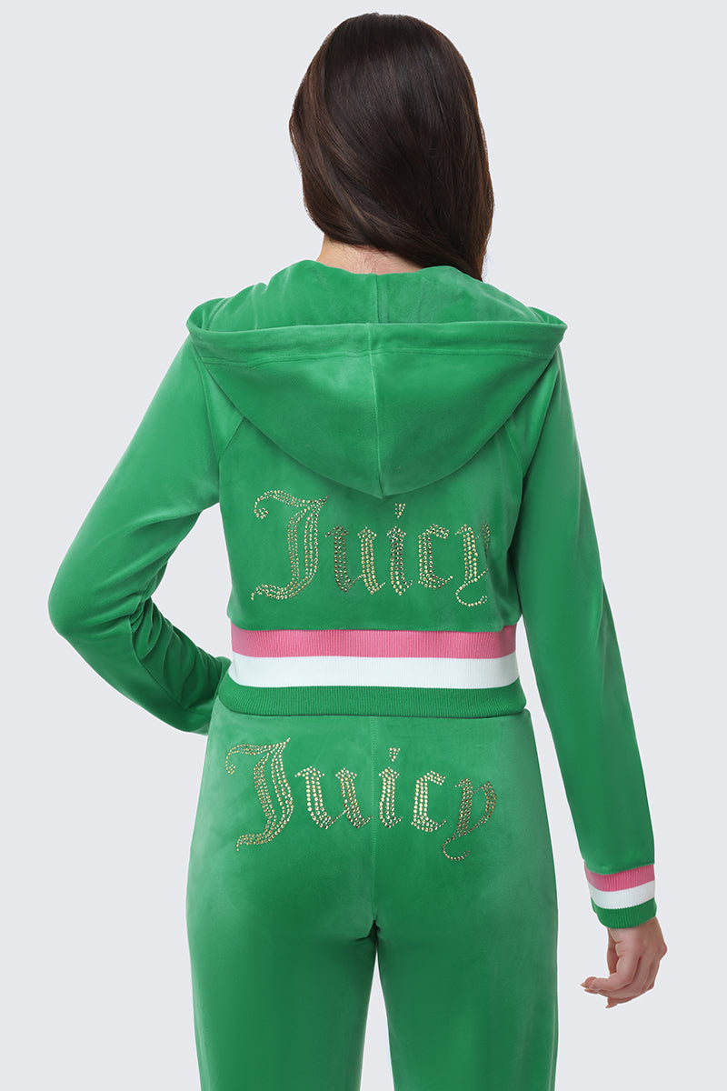 A woman in a green Heritage Stripe Bling Velour Zip Hoodie and matching pants faces away from the camera. Both pieces display a gold rhinestone "Juicy" logo, and the hooded jacket features pink and white striped waistband and cuffs.