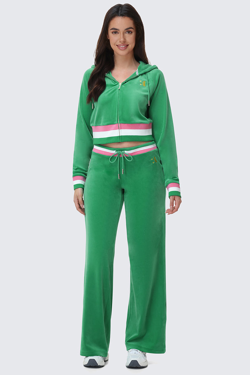 A woman faces forward wearing the Heritage Stripe Bling Velour Zip Hoodie by Juicy Couture in green with a rhinestone logo, white and red striped waistband and cuffs, matching wide-leg pants, long dark hair, and white sneakers.