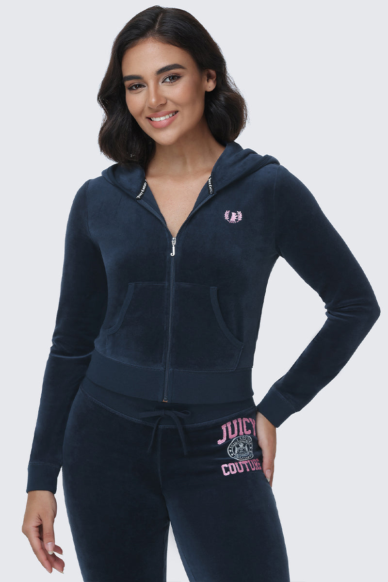 A woman stands and smiles with one hand on her hip against a plain background, wearing the Scottie Cotton Velour Hoodie—a navy blue, collegiate-inspired zip-up hoodie featuring the Scottie crest—paired with matching Juicy Couture text pants.