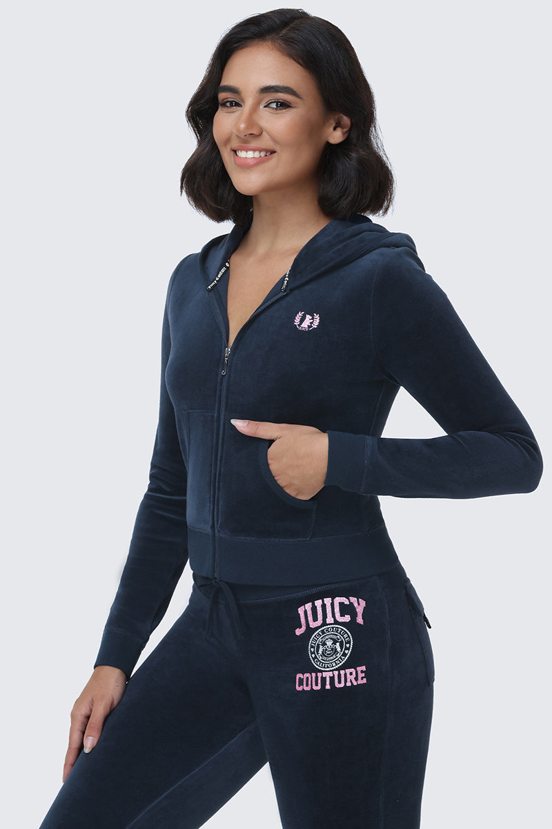 A woman smiles, wearing the Scottie Cotton Velour Hoodie in navy blue, featuring pink Juicy logo text and the signature Scottie crest. She stands against a light gray background with one hand in her hoodie pocket.