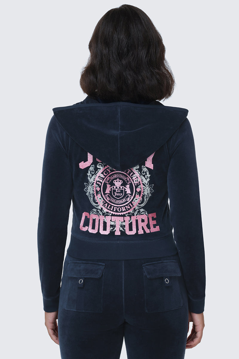 A woman seen from the back wears the Scottie Cotton Velour Hoodie in navy blue, featuring Juicy Couture and a pink Scottie crest logo on the back, giving her look a collegiate-inspired flair.