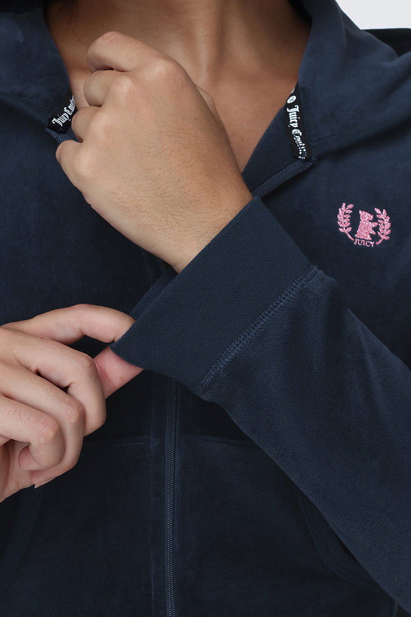 Wearing the Scottie Cotton Velour Hoodie, featuring a navy blue zip-up design, a pink Scottie crest logo, and "Juicy" embroidered on the chest, someone pulls at the sleeve to show off its collegiate-inspired style.