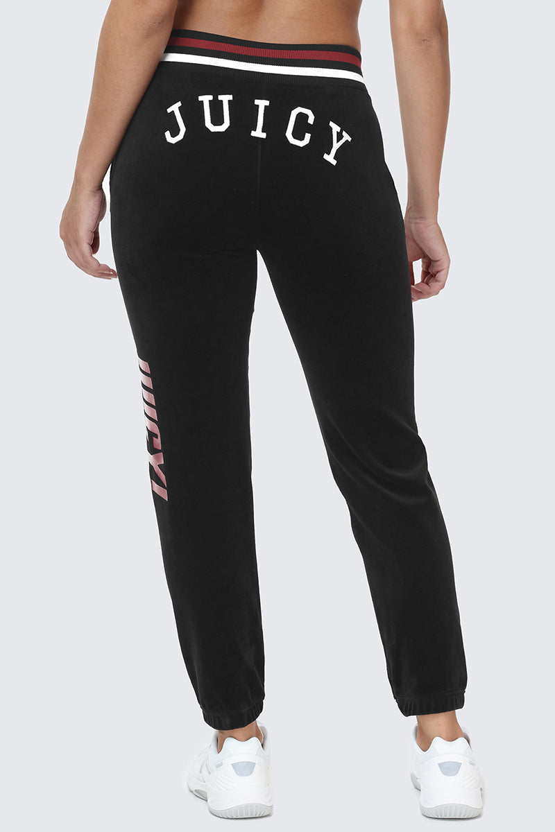 A person shown from behind wearing the Mixed Logo Cotton Velour Jogger in black with bold white "JUICY" lettering on the back, paired with white sneakers—a true sporty staple.