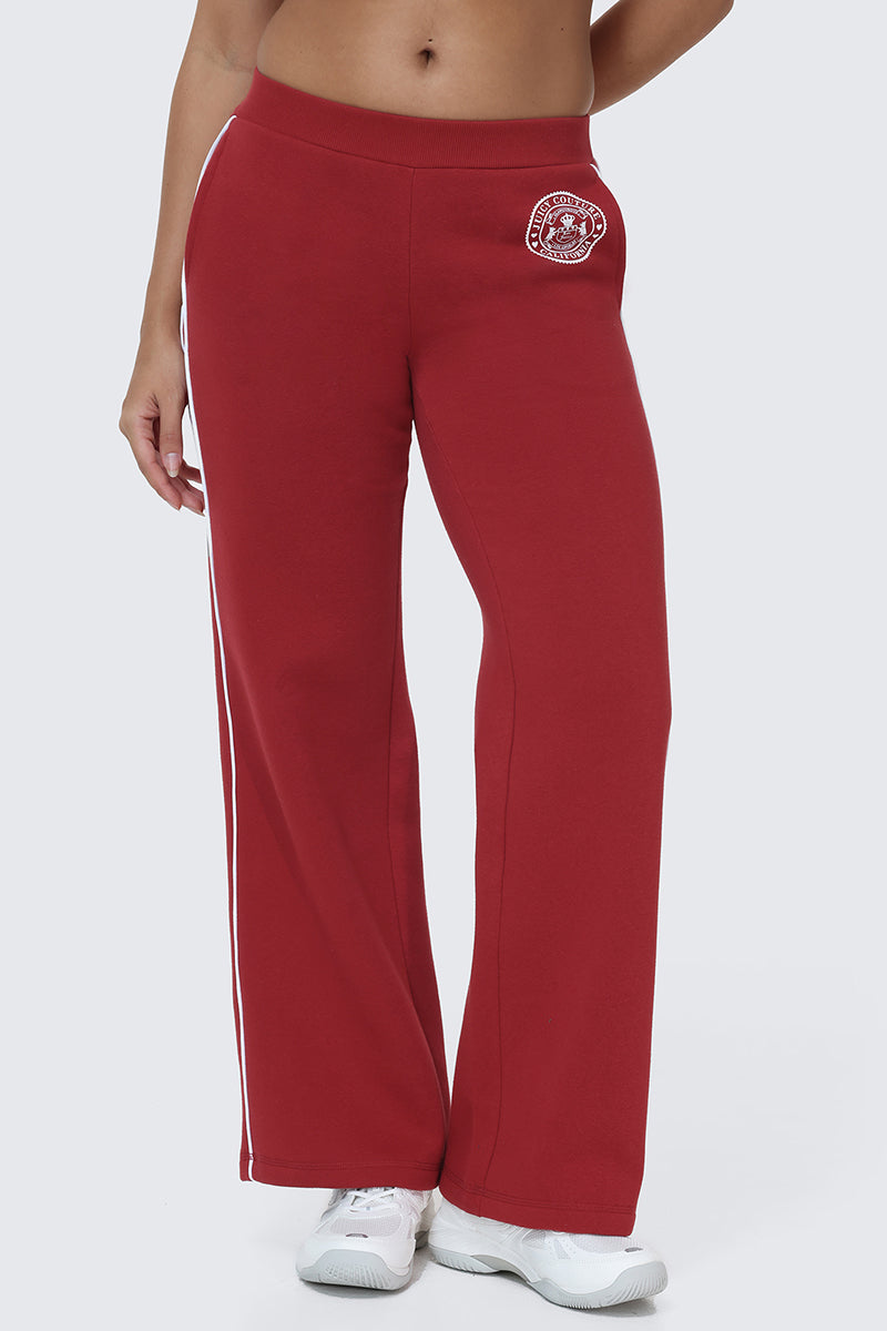 A person wearing the Scottie Fleece Wide Leg Pants—high-waisted red sweatpants featuring a small Scottie crest patch on the upper left thigh and white side stripes—paired with white athletic shoes. Only the lower torso and legs are visible.