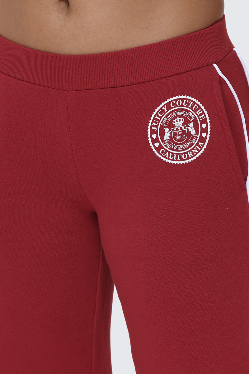 Close-up of a person wearing red Scottie Fleece Wide Leg Pants by Juicy Couture, featuring a white logo patch on the hip that reads "Juicy Couture California" around a crest design, giving these sweatpants a collegiate-inspired look.