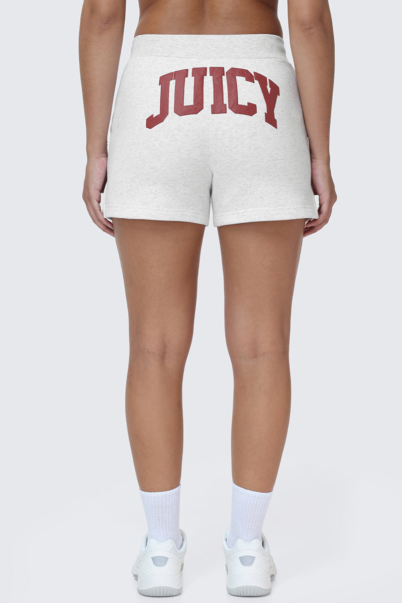 A person wearing the Scottie Fleece Booty Shorts, featuring the Juicy logo in bold red collegiate lettering on the back, paired with white socks and sneakers, standing against a plain background.