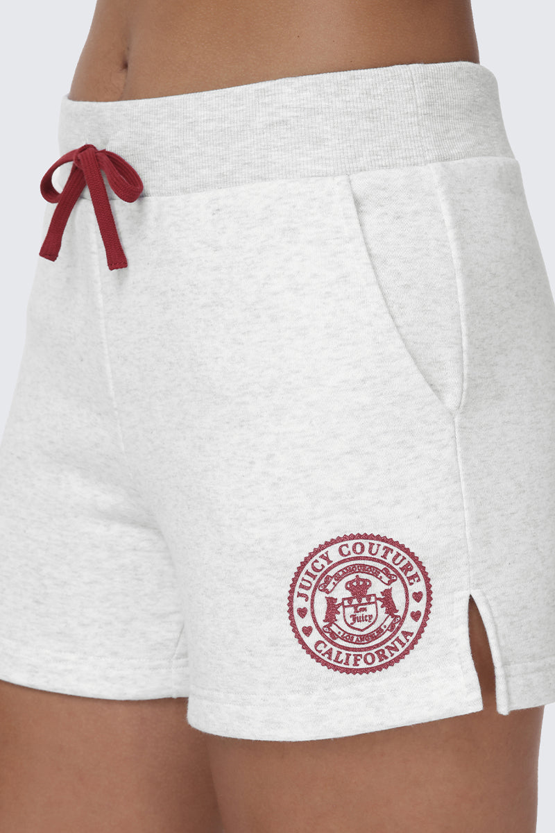 Close-up of a person wearing the Scottie Fleece Booty Shorts by Juicy Couture in light gray, featuring a red drawstring, collegiate-style Juicy Couture California logo patch near the bottom hem, side pockets, and a small slit.