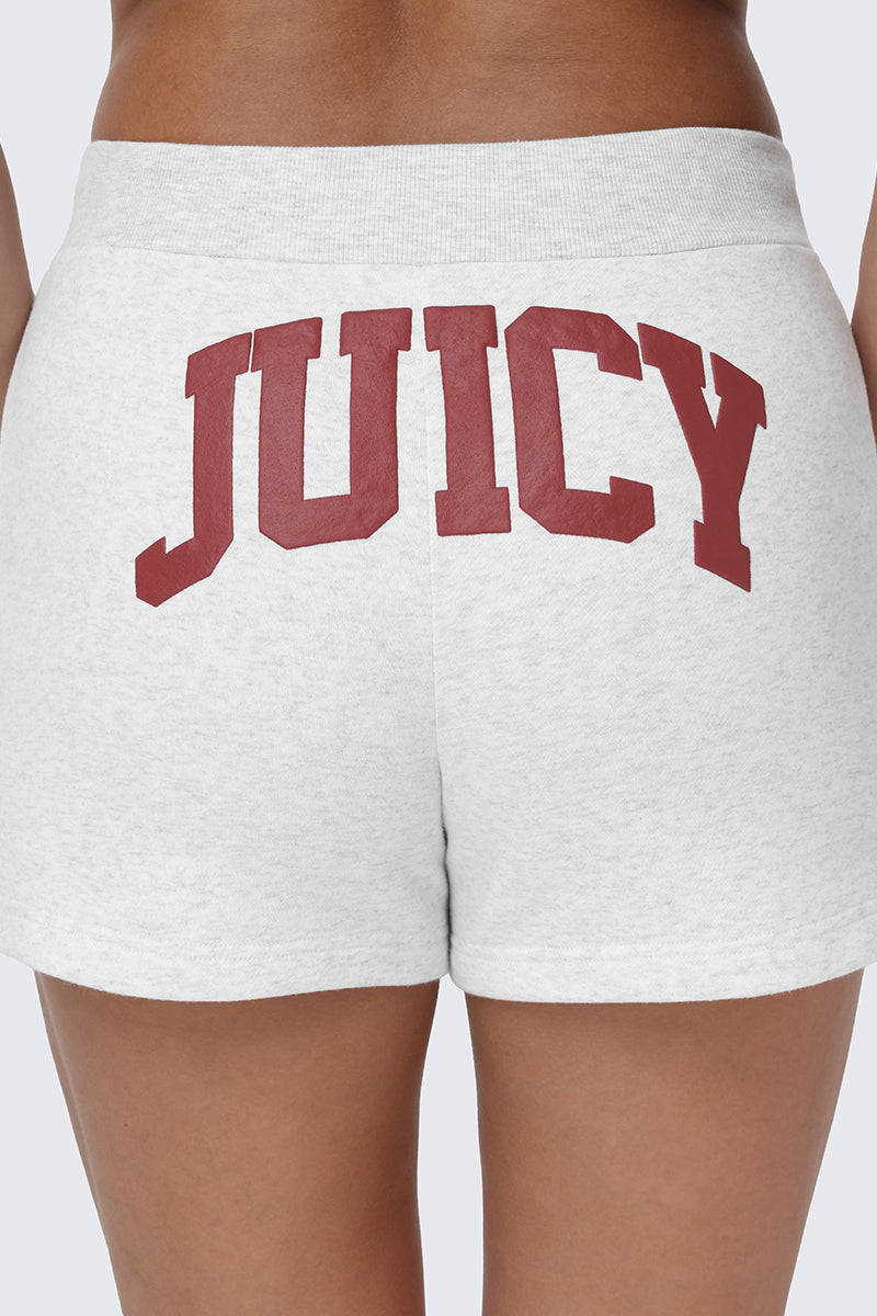 A close-up of a person wearing the Scottie Fleece Booty Shorts in light gray, featuring the Juicy logo printed in large red collegiate-style letters across the back.