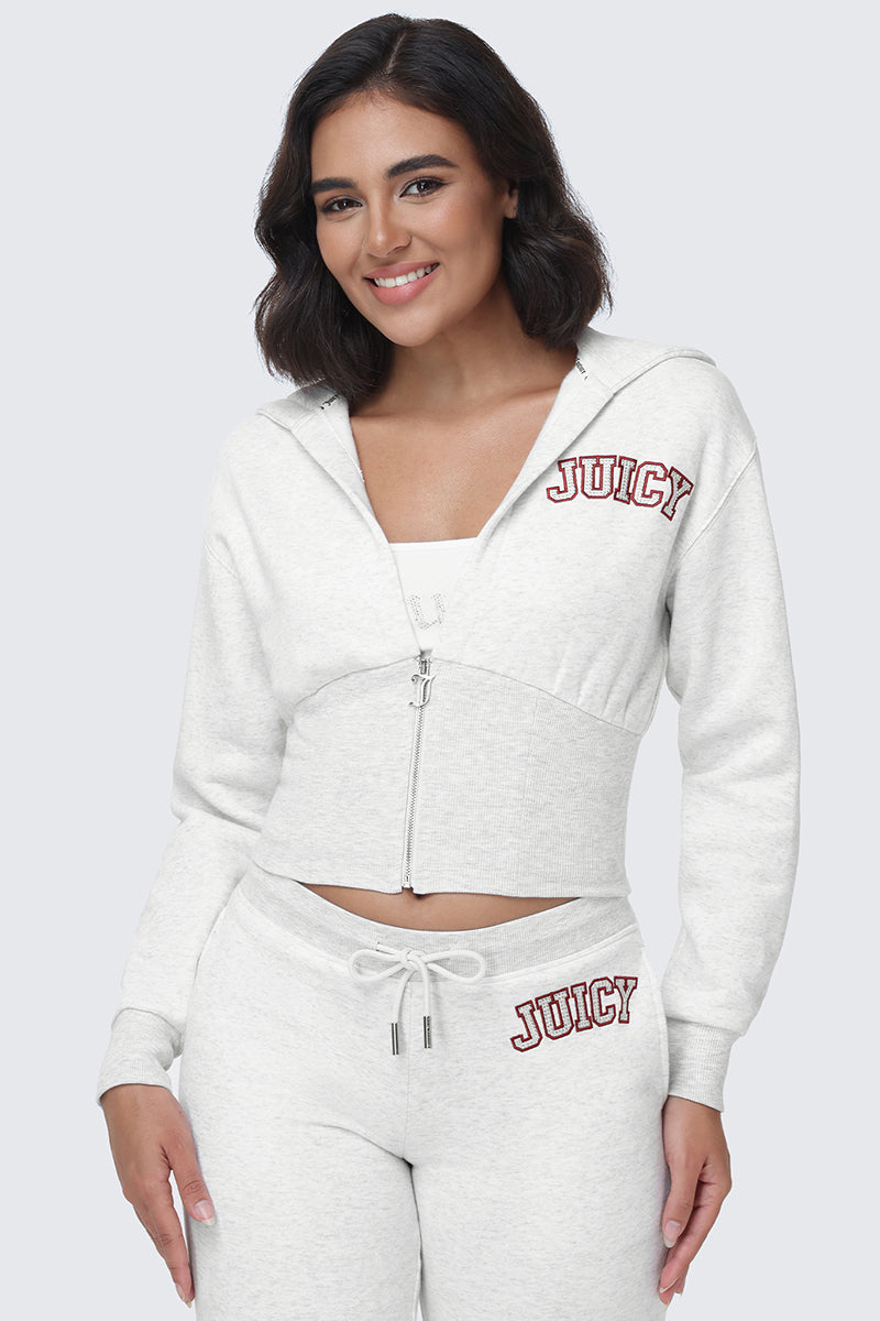A woman stands and smiles at the camera against a plain light background, wearing a light gray Small Bling Fleece Quarter Zip Hoodie from Juicy Couture with matching sweatpants featuring JUICY lettering. The fitted silhouette adds a modern touch to this iconic lounge look.