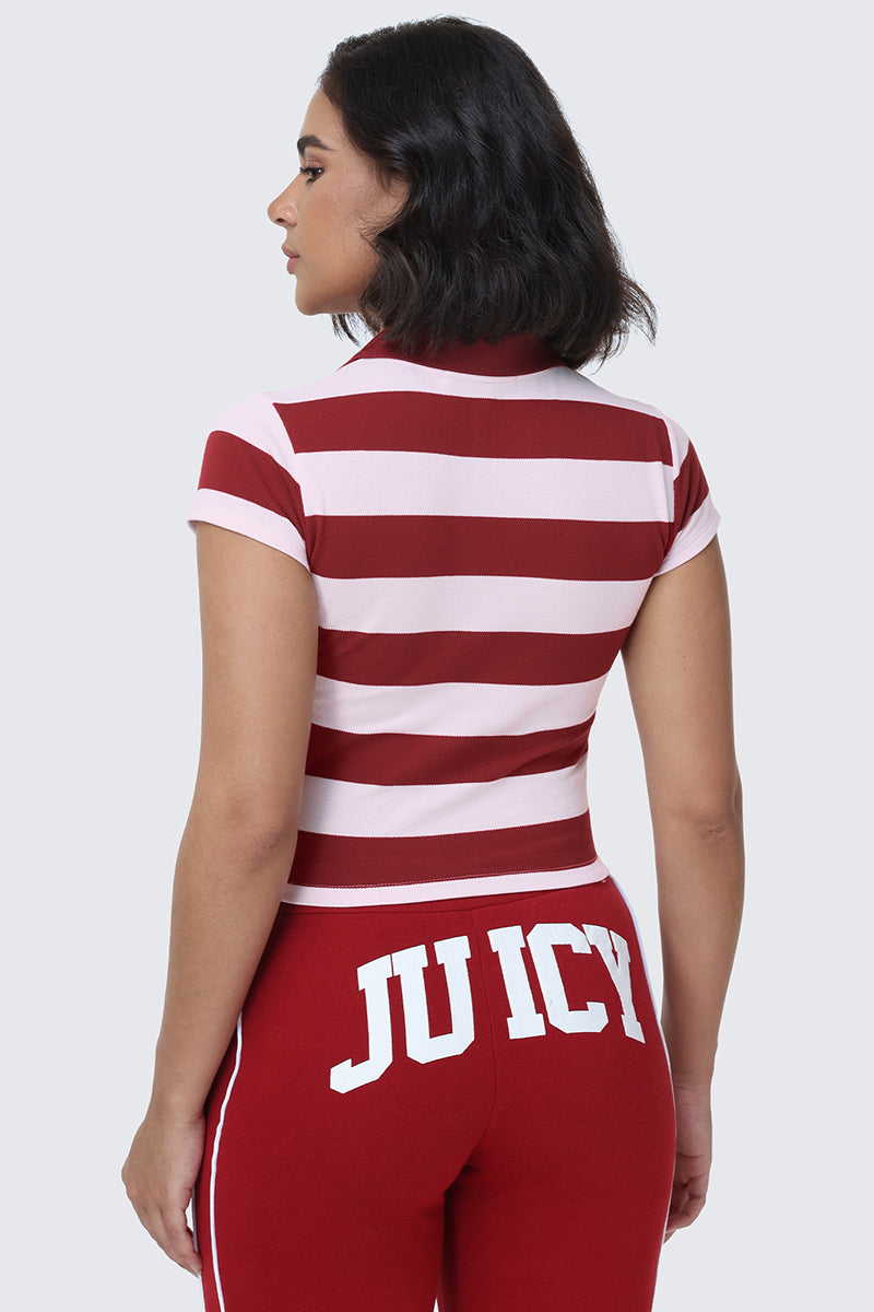 A woman with dark hair wears the Stripe Scottie Baby Polo, a red and pink striped cropped top, paired with red pants featuring “JUICY” in large white letters across the back. She is facing away from the camera against a light gray background.
