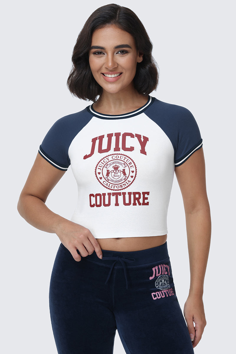 A woman with dark shoulder-length hair smiles while wearing the Scottie Baby Tee by Juicy Couture, a short-sleeve white and navy top with collegiate graphics, paired with matching navy pants featuring the iconic "Juicy Couture" text. She stands against a plain light background.