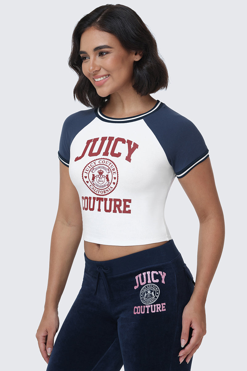 A woman with shoulder-length dark hair stands and smiles against a light gray background wearing the navy and white Scottie Baby Tee by Juicy Couture, featuring collegiate graphics and signature logos, paired with matching navy sweatpants.