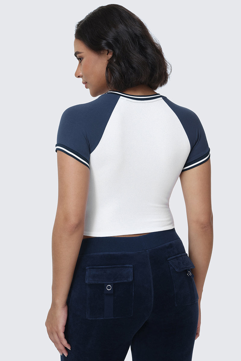 A woman is shown from behind, wearing the Scottie Baby Tee—a white short-sleeved ringer top with navy blue raglan sleeves and trim—paired with dark blue Juicy Couture velour cargo pants, set against a light grey background.