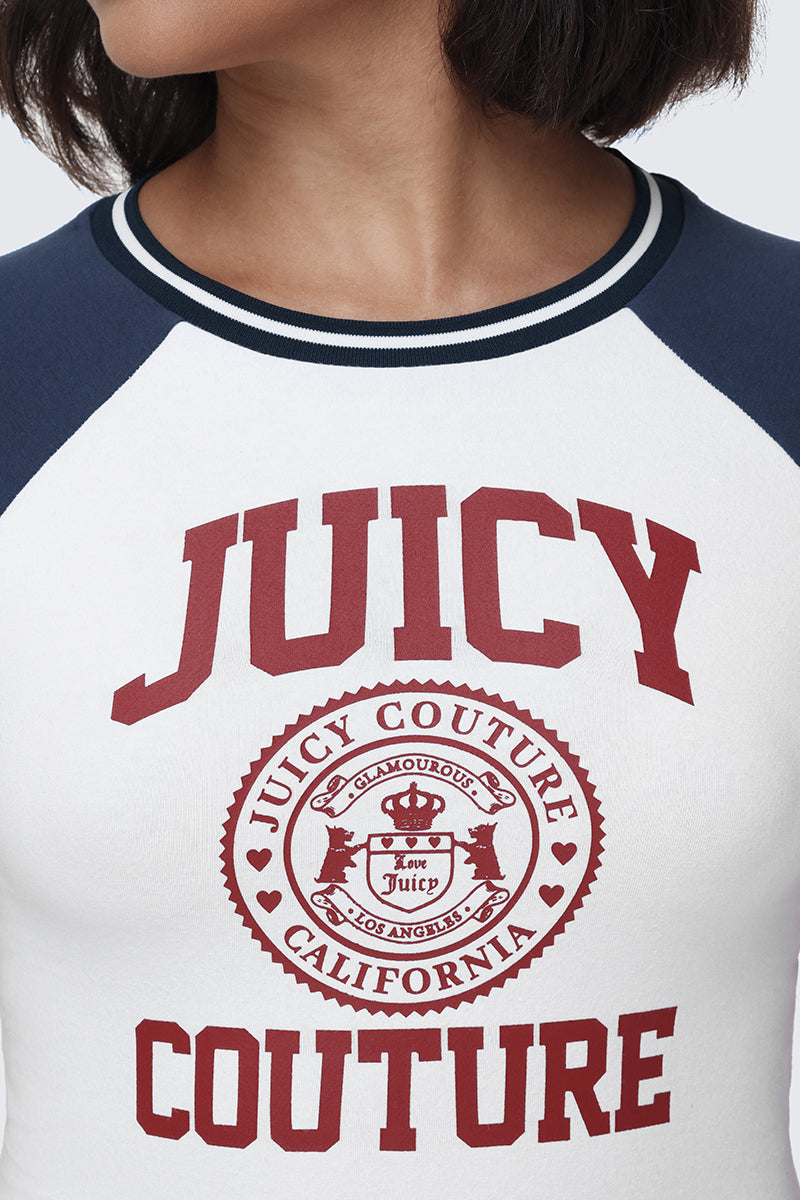 A person is wearing the Scottie Baby Tee, a white top with navy sleeves featuring JUICY COUTURE in bold red letters and a circular crest logo that reads "Juicy Couture California Los Angeles" on the front, showcasing classic collegiate graphic style.