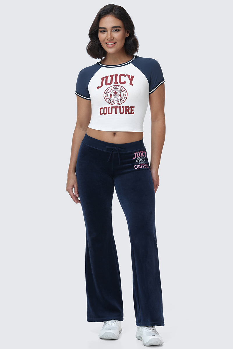 A woman stands and smiles, wearing the Scottie Baby Tee by Juicy Couture, which features white and navy coloring with collegiate graphics, paired with navy blue velour pants displaying the pink Juicy Couture logo on the hip and white sneakers, set against a plain light background.