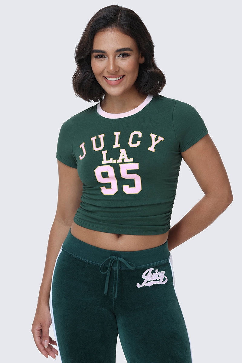 A woman with short dark hair smiles as she wears the L.A. Graphic Baby Tee in green, featuring varsity-style JUICY L.A. 95 graphics in bold pink and yellow, paired with coordinating green sweatpants adorned with the Juicy logo on the waistband.