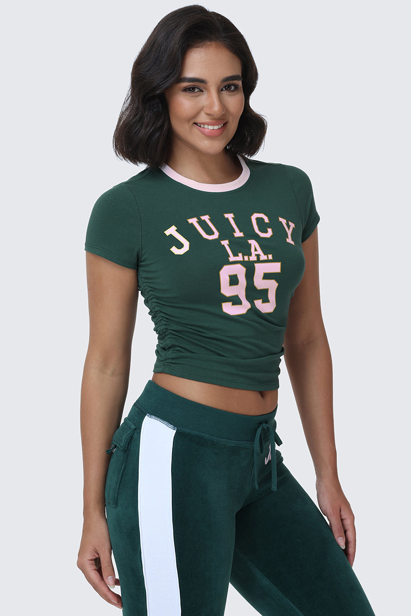 A woman smiles at the camera wearing the L.A. Graphic Baby Tee, a dark green fitted top featuring bold varsity-style JUICY L.A. 95 graphics in pink, paired with matching green pants that have white side stripes.