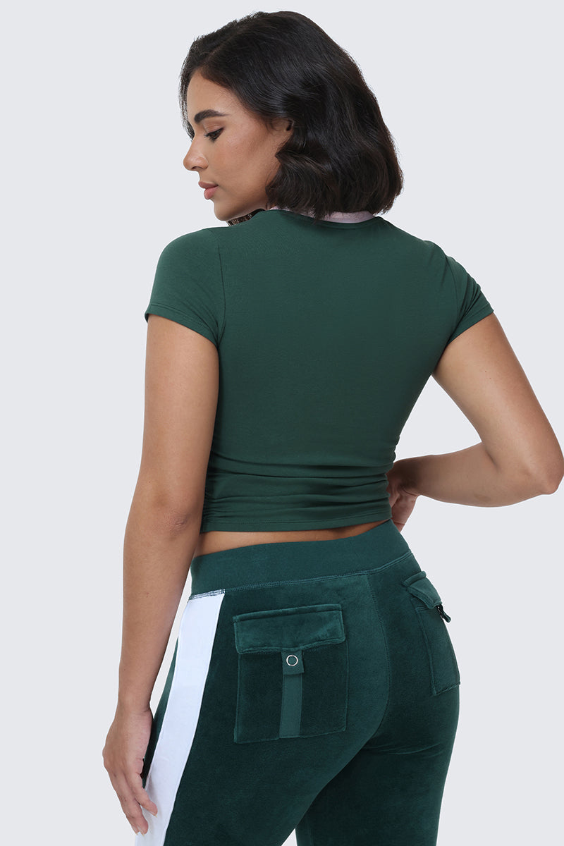 A woman with short dark hair is wearing the L.A. Graphic Baby Tee, a fitted green crop top inspired by Juicy L.A. 95, paired with green pants featuring white side stripes and back pockets, as she stands with her back to the camera.