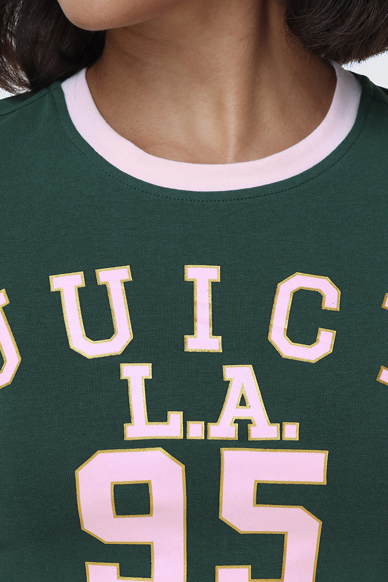Close-up of a person wearing the L.A. Graphic Baby Tee, a dark green fitted top featuring large pink and yellow "JUICY L.A. 95" varsity graphics across the chest. Only the lower face and neck are visible.