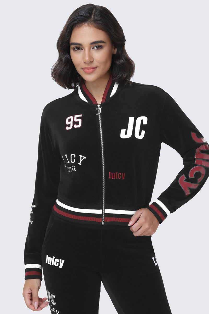 A woman stands against a plain light background wearing the Mixed Logo Cotton Velour Bomber Jacket by Juicy Couture, featuring black cotton velour fabric, white and red Juicy graphics, a zip-front closure, and striped cuffs and collar.