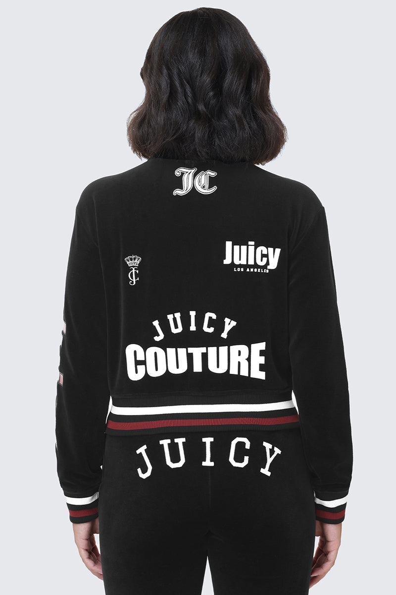 A person with shoulder-length dark hair is seen from the back wearing the Mixed Logo Cotton Velour Bomber Jacket by Juicy Couture. The black cotton velour bomber features multiple white Juicy Couture logos and graphics on both the jacket and pants, giving it a sporty throwback vibe.