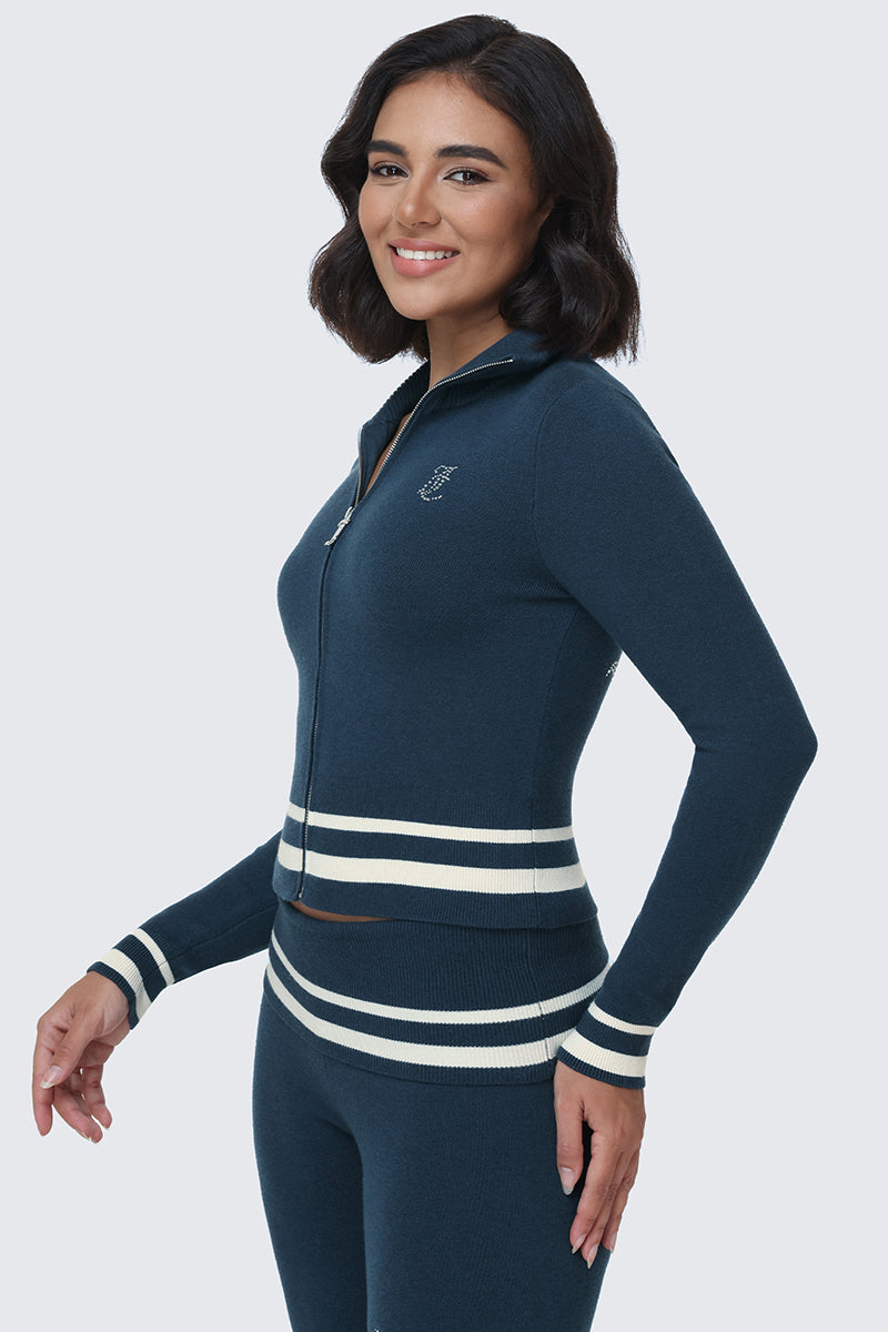 A woman stands sideways and smiles at the camera against a plain light background, wearing the Big Bling Knit Jacket in navy blue with matching pants—both featuring white stripes on the cuffs and waist and a sparkling rhinestone Juicy logo on the back.