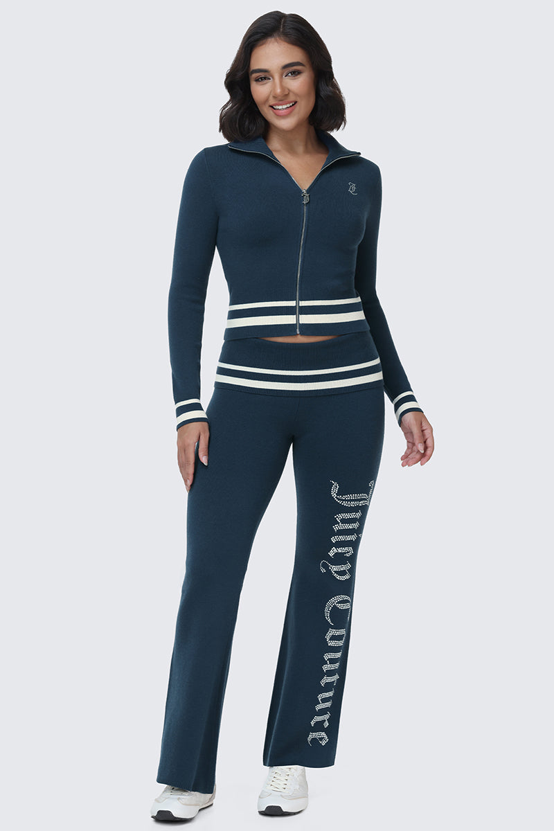 A woman stands and smiles against a plain light background, wearing the Big Bling Knit Jacket in navy blue paired with matching flared pants featuring white stripes and the Juicy logo spelled out in rhinestones on one leg.