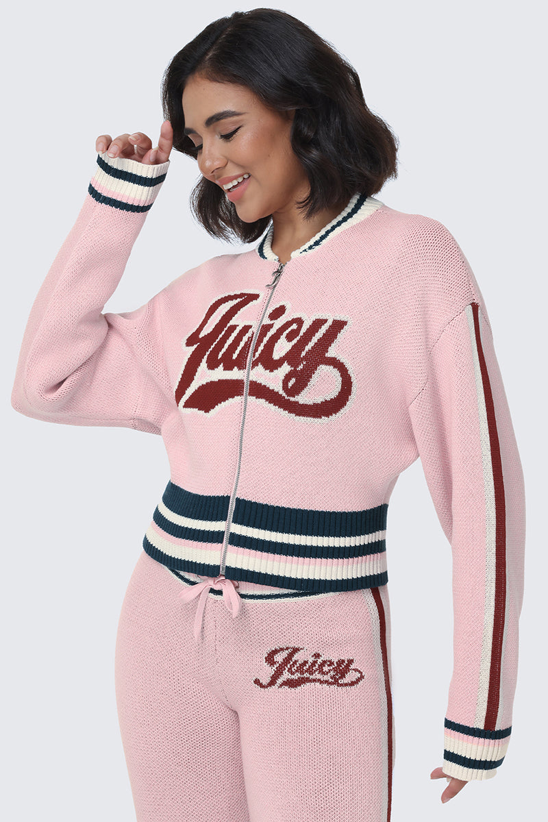 A woman smiles while wearing the Contrast Knit Bomber Jacket, a pink knit zip-up sweater with matching pants featuring the Juicy script logo on both. The outfit is accented with navy and white striped trim on the collar, cuffs, waistband, and pants, giving it a playful varsity jacket vibe.
