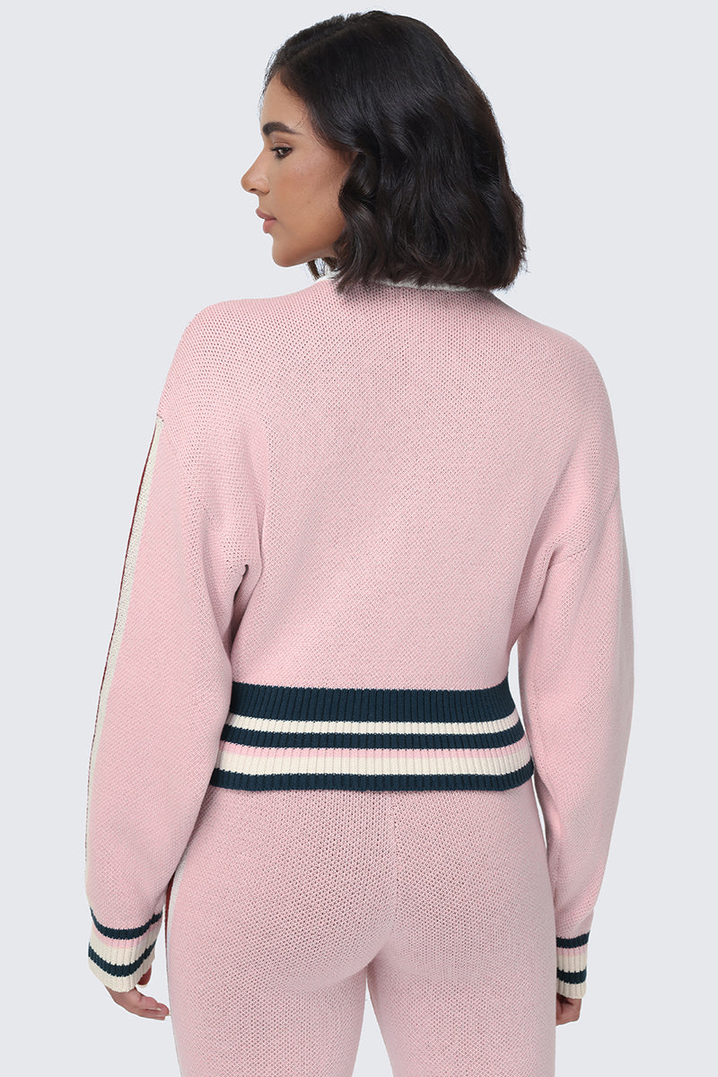 A woman with short dark hair, seen from behind, wears the Contrast Knit Bomber Jacket in light pink, paired with matching pants. The jacket features wide navy and white striped cuffs and waistband inspired by classic varsity styles. The background is plain light gray.