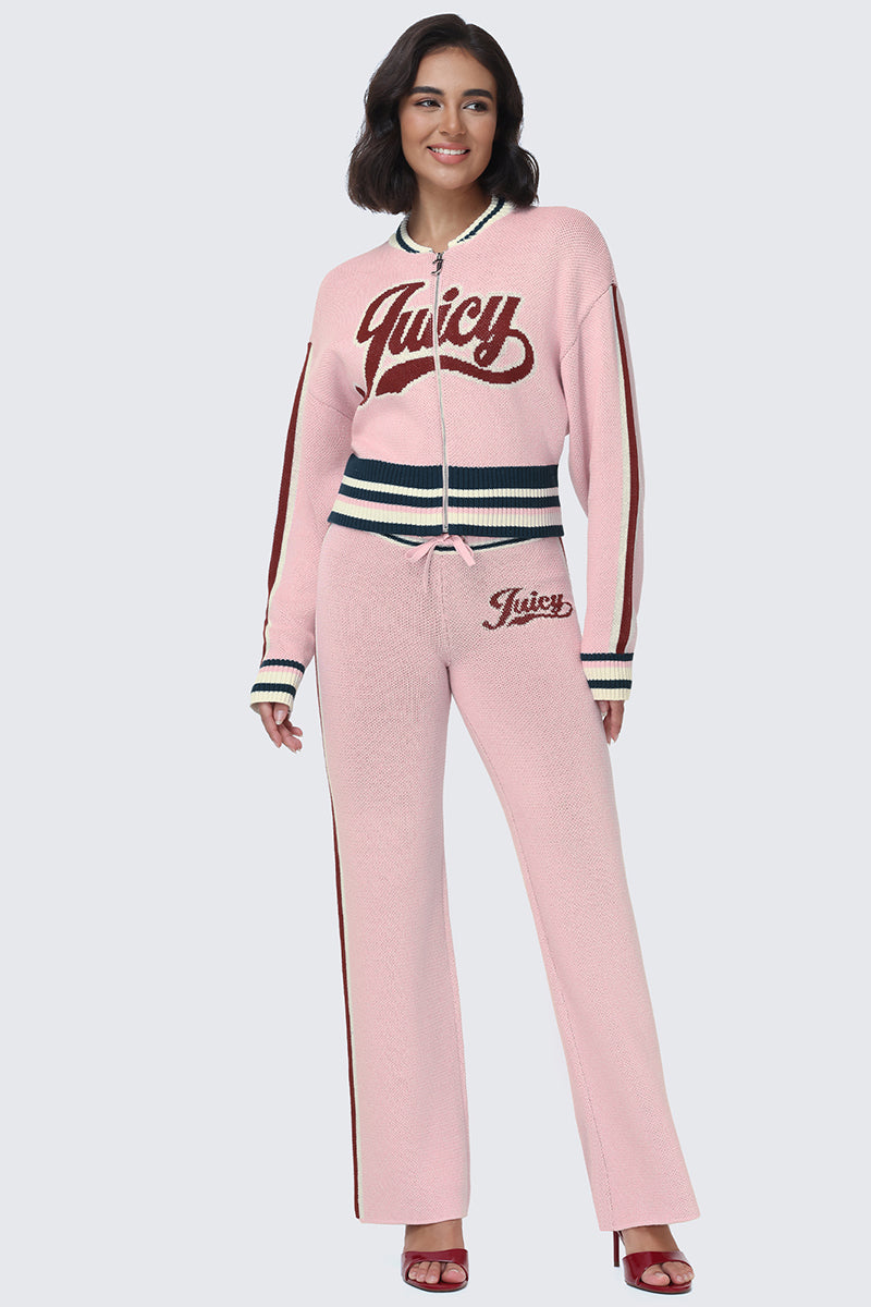 A woman stands and smiles against a plain background, wearing the Contrast Knit Bomber Jacket in light pink with striped varsity-style trim, paired with matching pants featuring the Juicy script logo and red high heels.