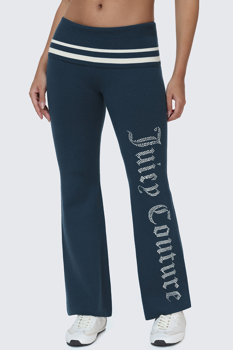 A person wearing the Juicy Couture Big Bling Knit Flare Pants in dark blue, featuring white waistband stripes and the sparkling Juicy Couture logo in rhinestones down the leg, paired with white sneakers.